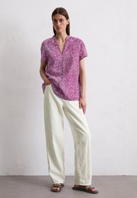 Kurzärmlige, rosa-weiß gemusterte Bluse mit V-Ausschnitt, kombiniert mit hellen, locker sitzenden Hosen und dunklen Sandalen.