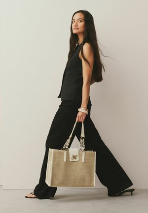 Bēša austā rokassoma ar baltām detaļām, ar ķēdes siksnām un zeltainu logotipu uz metāla detaļām. Soma ir saskaņota ar melnu apģērbu.