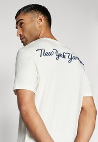 Balta kokvilnas T-krekls ar tumši zilu izšūtu "New York Yankees" tekstu uz muguras. Īsas piedurknes un noapaļots kakls.