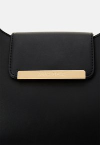 Schwarze Lederhandtasche mit einer goldenen Metallplakette, auf der "ANNA FIELD" eingeprägt ist. Glatte Textur und Klappverschluss-Design.