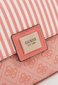 Gros plan d’un tissu rayé rose et blanc avec une plaque métallique du logo GUESS sur une surface texturée rose de sac.