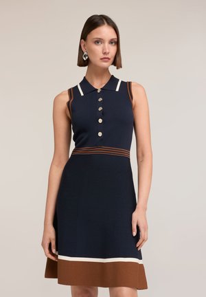 Kobieta ubrana w granatową sukienkę bez rękawów z brązowymi i białymi wykończeniami, guzikami z przodu oraz geometryczne srebrne kolczyki, stojąca na neutralnym tle.