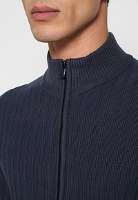 Donkerblauwe geribbelde trui met een hoge kraag en een ritssluiting. De stof is zacht en gebreid, met verticale lijnen en een gestroomlijnd ontwerp.