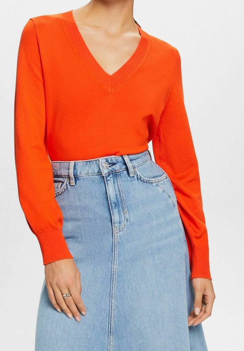 Femme portant un pull à col en V manches longues de couleur orange vif, rentré dans une jupe en jean bleu clair taille haute avec des poches avant.