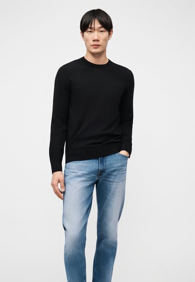 Άνδρας όρθιος, φοράει μαύρο πουλόβερ μακρυμάνικο και ανοιχτόχρωμο μπλε τζιν, με το ένα χέρι στην τσέπη, μπροστά σε απλό ανοιχτόχρωμο φόντο.