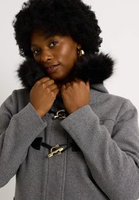 Abrigo de lana gris con borde de piel negra, que presenta un cierre de toggles de hardware dorado y un corte relajado. La textura parece suave y cálida.