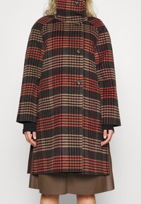 Mujer que lleva un abrigo de cuadros hasta la rodilla en tonos rojos, marrones y beiges con botones negros y un cuello alto, sobre una falda marrón.