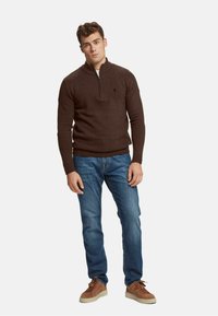 Suéter marrón de punto con cuello de media cremallera, combinado con jeans de mezclilla azules y zapatillas de ante marrones, llevado por un modelo masculino de pie.