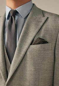 Grau karierter Blazer mit einer taillierten Form, kombiniert mit einem hellblauen strukturierten Hemd und einer dunkelgrauen Krawatte. Mit einem braunen Einstecktuch in der Brusttasche.