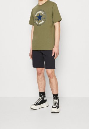 Mand iført en olivengrøn Converse All Star t-shirt, sorte shorts og sorte Converse højtop sneakers med hvide såler og snørebånd.