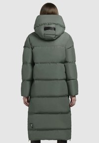 Parka lunga e voluminoso in verde spento, con un grande cappuccio, cuciture orizzontali e una toppa con logo sulla schiena. Tessuto liscio.