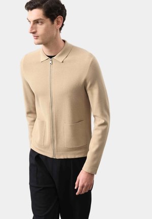 Cárdigan beige con cremallera, cuello, dos bolsillos delanteros y diseño ajustado. Confeccionado con un material de punto fino que presenta una textura suave.