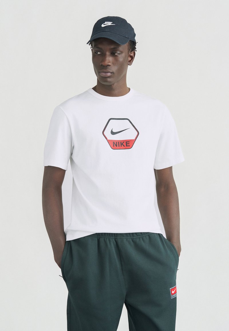 Νεαρός άνδρας που φοράει λευκό μπλουζάκι Nike, σκούρο πράσινο παντελόνι με μπαλώματα και σκούφο Nike, στέκεται με τα χέρια στις τσέπες.