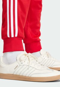 Biele kožené tenisky s podrážkou z gumy, ozdobené tromi modrými pruhmi a štítkom "SAMBA". Kombinované s červenými teplákmi s bielymi pruhmi.