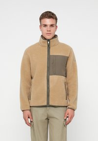 Junger Mann in einer beigen Fleecejacke mit dunklem grünen Reißverschluss-Brusttasche und passenden Cargo-Hosen, der vor einem weißen Hintergrund steht.