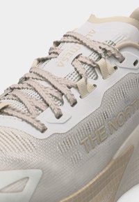 Κοντινό πλάνο ενός μπεζ και λευκού αθλητικού παπουτσιού με υφασμένα κορδόνια και τη σήμανση "THE NORTH" στο πλάι.