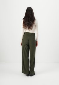 Pantalons larges verts avec des côtes verticales, taille haute et coupe décontractée. Portés avec un haut à manches longues crème et des chaussures noires.