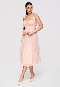 Femme aux cheveux foncés portant une robe corset rose pâle avec des bretelles détaillées et une jupe en tulle à pois, chaussée de chaussures crème à talons hauts avec bride à la cheville.