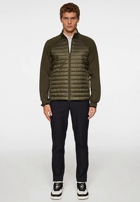 Oliivinvihreä tikattu takki, jossa on täysi vetoketju, mustat hihat ja kevyt malli. Yhdistetty tummiin housuihin ja kaksivärisiin tennareihin.