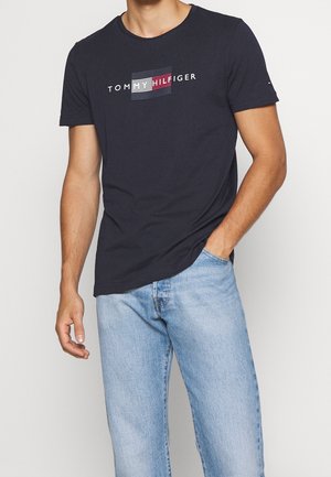 Mężczyzna w granatowej koszulce Tommy Hilfiger z logo i jasnoniebieskich dżinsach, jedna ręka w kieszeni, na jednolitym tle.
