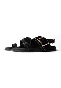 Paar schwarze flache Sandalen mit breiten Riemen, einer mit großer goldener Schnalle und verstellbarem Knöchelriemen, auf weißem Hintergrund.