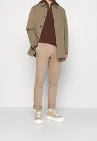 Person klädd i ljusbruna uppvikta byxor, beige sneakers, en brun zip-up tröja och en olivgrön knappad kappa stående mot en enfärgad bakgrund.