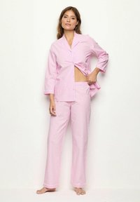 Roze pyjama set met een knoopsluiting bovenstuk met driekwart mouwen, een borstzak en bijpassende broek. Zacht materiaal met kleine witte stippen.