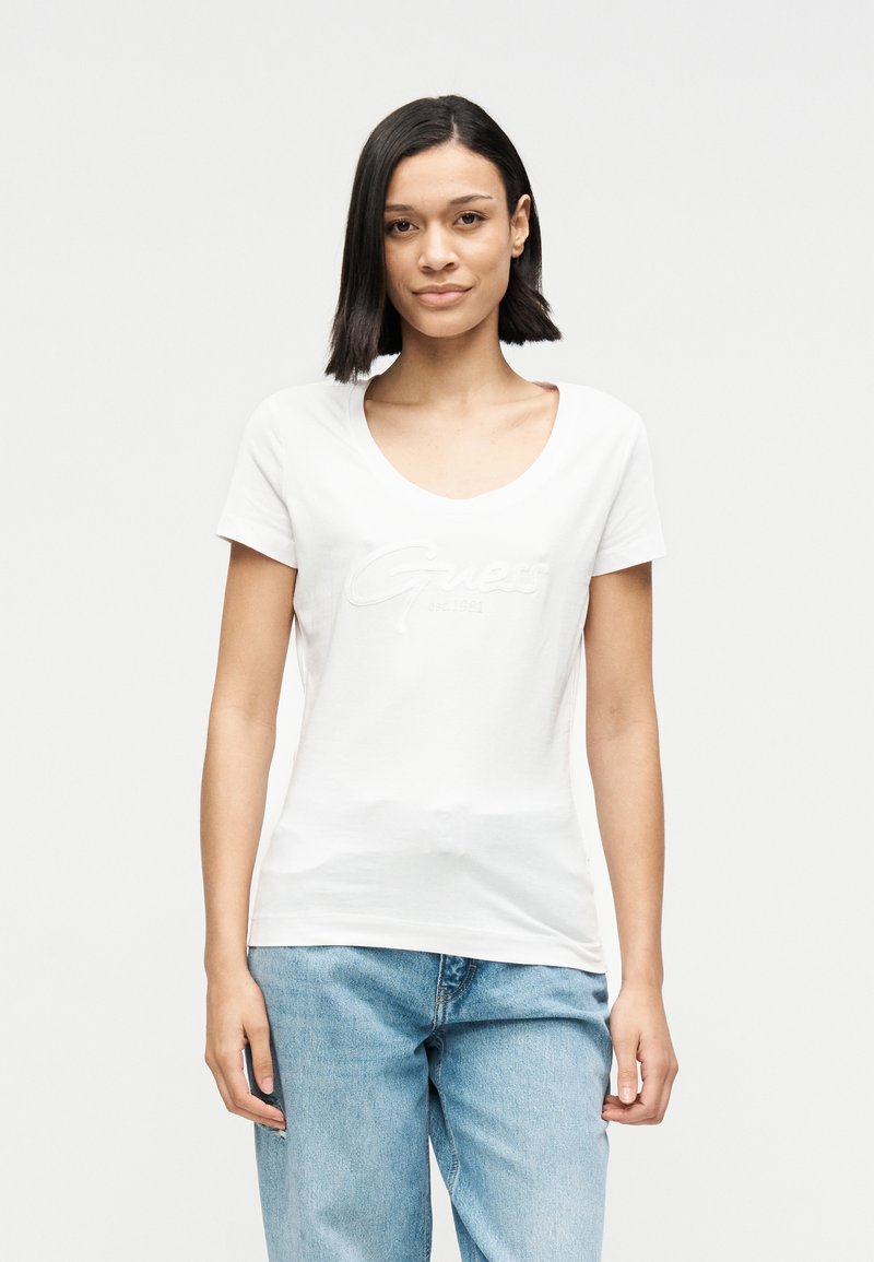 Λευκό βαμβακερό T-shirt με V-λαιμόκοψη και κοντά μανίκια. Διαθέτει διακριτικό τονικό λογότυπο "Guess" στο στήθος. Συνδυάζεται με ανοιχτόγαλαντζες τζιν.
