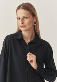 Mustpandanülik särk, millel on krae, sujuv tekstuur. Modell kannab suurt, musta ja kuldset mustriga kõrvarõngast. Juuksed on sirged ja jaotatud.