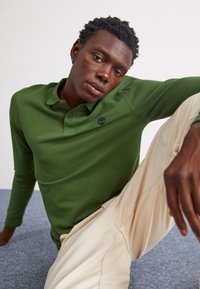 Polo de mangas longas verde escuro com colarinho de dois botões e um pequeno logo no peito, combinado com calças beje claras. Tecido suave e liso.