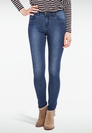 Skinny jeans in dark blue denim, featuring a high waist, two front pockets, and subtle stitching details. Paired with beige ankle boots.