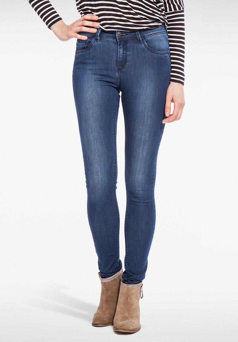 Jeans ajustados en denim azul oscuro, con cintura alta, dos bolsillos frontales y detalles de costura sutiles. Combinados con botas de tobillo beige.