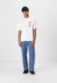 Hvid grafisk t-shirt med farverig "SR" print og tekst på fronten, parret med lyseblå jeans og sorte sko med gummisål.