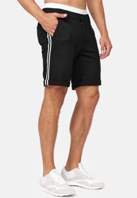 Schwarze Shorts mit umgeschlagenen Säumen, weißen Seitenstreifen, Knopf- und Reißverschluss, sowie zwei Fronttaschen. Hergestellt aus weichem, atmungsaktivem Stoff.