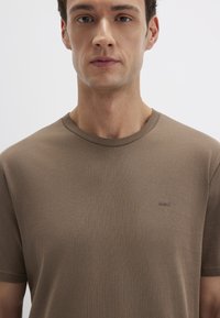 Braun farbiges Baumwoll-T-Shirt mit Rundhalsausschnitt und kurzen Ärmeln. Verfügt über eine dezente Rippstruktur und ein kleines Logo auf der linken Brustseite.