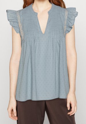 Blusa azzurro chiaro con maniche a sbuffo, scollatura a V profonda e motivo a pois testurizzato. Presenta pieghe verticali e dettagli in pizzo.