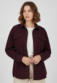 Burgundy corduroy shirt jacket with a collared neckline, two buttoned chest pockets, and a relaxed fit, paired with a white top and beige pants.