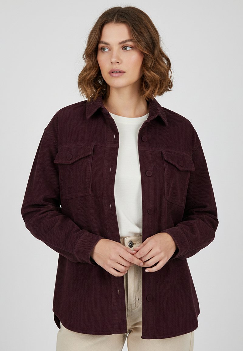 Burgundy corduroy shirt jacket with a collared neckline, two buttoned chest pockets, and a relaxed fit, paired with a white top and beige pants.