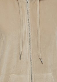 Veste en tissu doux beige avec fermeture éclair argentée à l'avant et cordons assortis avec embouts métalliques argentés.