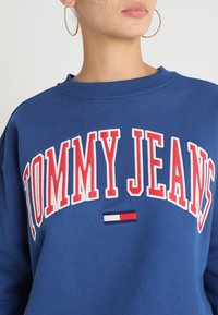 Blå tröja med "TOMMY JEANS" i stora röda bokstäver. Har en liten logotypdetalj i röd, vit och marinblå längst ner. Mjuk textur.