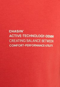 Tessuto rosso con superficie liscia che presenta testo bianco: "CHASIN' ACTIVE-TECHNOLOGY-DENIM CREA UN EQUILIBRIO TRA COMFORT-PERFORMANCE-UTILITY".