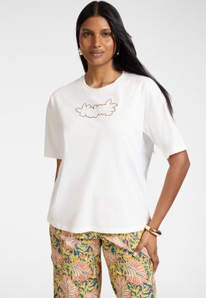 Vrouw met lang zwart haar die een witte top met korte mouwen en bloemenuitsnede draagt, met kleurrijke, patroonbroeken, staand met één hand op haar heup.