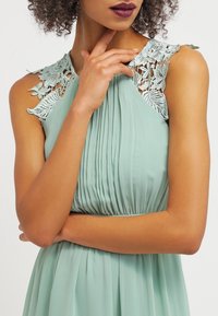 Mint vihreä mekko, jossa on poimutuksia ja monimutkaisia pitsikoristeita hartioilla. Kevyt, helposti valuva kangas, jossa on istuva yläosa.
