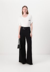 Witte t-shirt met "HUGO" logo, hoge zwarte broek met een wijde pijp en riem, zwarte handtas en zwarte enkellaarzen.