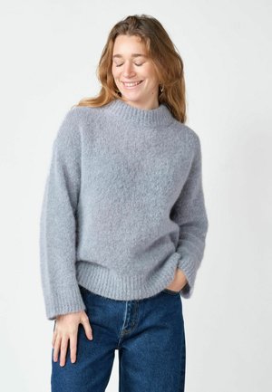 Femme aux yeux fermés souriant, portant un pull gris clair pelucheux et un jean bleu, debout avec une main dans la poche sur fond blanc.