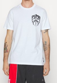 Hombre con una camiseta blanca de Puma con un logo de baloncesto en el pecho, pantalones cortos de baloncesto negros y rojos, y tatuajes visibles en los brazos.
