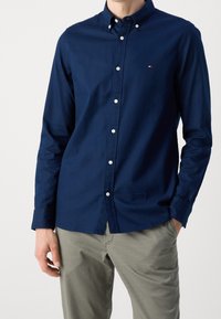 Chemise boutonnière bleu marine avec un col, des manches longues et des boutons blancs. Petit logo sur la poitrine. Portée avec un pantalon de couleur claire.