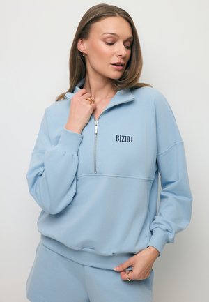 Vrouw draagt lichtblauwe zip-up sweater en bijpassende korte broek, houdt de kraag vast, kijkt iets naar beneden en opzij.