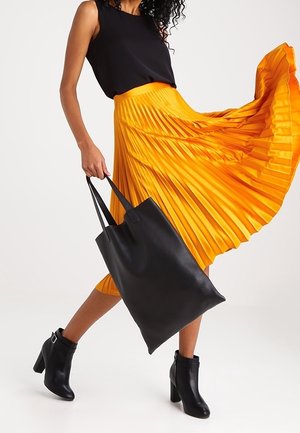 Zwarte mouwloze top, gele midi-rok met plooien, zwarte leren totebag en enkellaarzen. De rok heeft verticale plooien voor textuur.