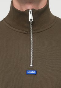 Πουλόβερ σε ελιά πράσινο με φερμουάρ στο λαιμό, από ριμπ ύφασμα. Διαθέτει ασημί φερμουάρ και μπλε λογότυπο "HUGO" στο στήθος. Ομαλή υφή.